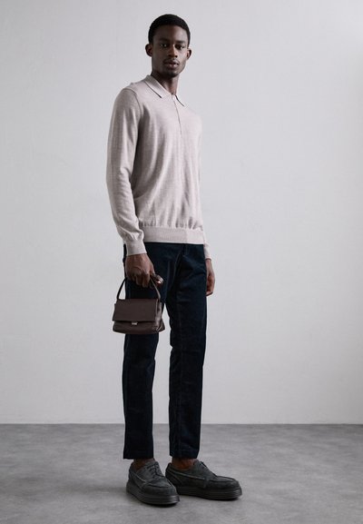Helles beige Langarm-Polo mit einem Reißverschlusskragen, kombiniert mit dunkelblauer Jeans und grauen Schuhen. Hält eine kleine dunkle Handtasche.