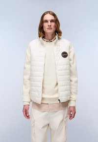 Helle, gesteppte Jacke mit hohem Kragen und besticktem Patch, getragen über einem cremefarbenen Pullover und beigen Cargo-Hosen.