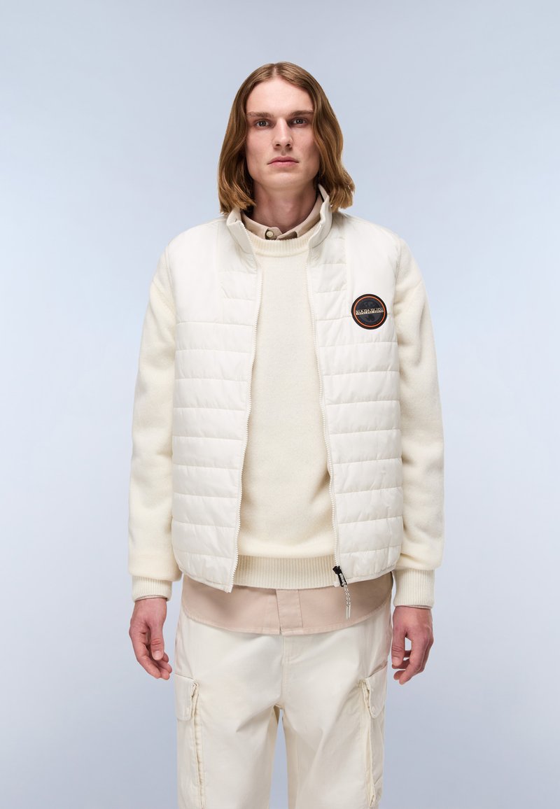 Helle, gesteppte Jacke mit hohem Kragen und besticktem Patch, getragen über einem cremefarbenen Pullover und beigen Cargo-Hosen.