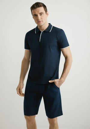 Młody mężczyzna w granatowej koszulce polo z krótkim rękawem z białą lamówką i dopasowanych granatowych krótkich spodenkach, pozujący z jedną ręką w kieszeni.