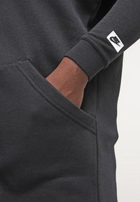 Tmavě šedá mikina Nike, rukáv se částečně nachází v přední kapse, na zápěstí je viditelné logo Nike.