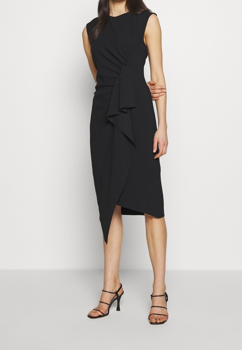 Femme portant une robe midi noire asymétrique sans manches avec un détail de volants drapés, associée à des sandales noires à talons hauts et à brides.