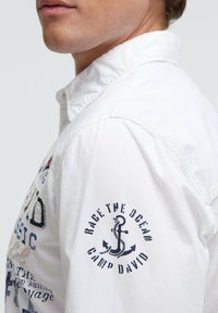 Weißes Denimhemd mit Knopfreißkragen, besticktem Text und Anker-Designs am linken Ärmel sowie sichtbaren Nahtdetails.