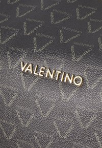 Μαύρο υφασμένο δέρμα με επαναλαμβανόμενο χρυσό μοτίβο "Valentino". Το όνομα της μάρκας είναι ευδιάκριτο σε χρυσή μεταλλική λεπτομέρεια.