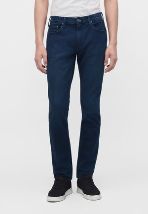 Personne portant un jean droit bleu foncé, une chemise blanche et des baskets noires à semelles blanches, debout devant un fond uni.
