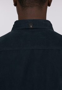 Camicia in corduroy scuro con colletto strutturato e una piccola etichetta nera sul retro. Il tessuto presenta delle righe verticali.