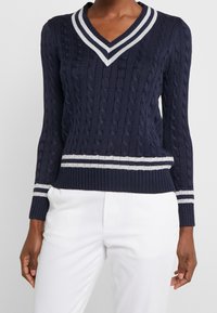 Granatowy sweter z splotem w warkocze, z dekoltem w kształcie V i ściągaczami na mankietach. Posiada białe paski wzdłuż dekoltu i dolnego wykończenia, oferując teksturowany design.