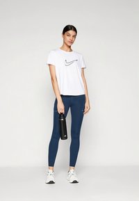 T-shirt branca de mangas curtas com logótipo preto, combinada com leggings azul-marinho. Modelo segura uma garrafa de água preta e calça ténis brancos.