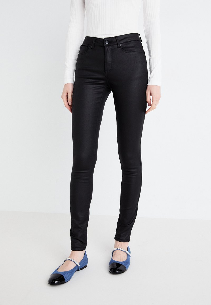 Black, slim-fit pants made of stretchy material, featuring a high waistband and five pockets. Paired with blue flats and a white ribbed top.