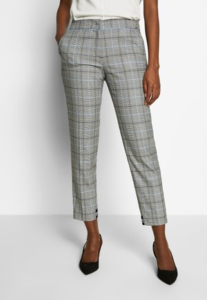 Frau trägt graue karierte, konisch geschnittene Hose mit hellblauen Linien, weißes Oberteil und schwarze High-Heels, steht vor weißem Hintergrund.