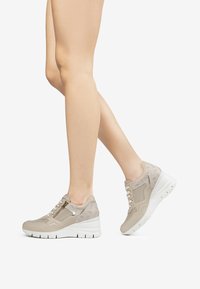 Zapatillas deportivas beige con parte superior de malla texturada, detalle de cremallera dorada y suela blanca gruesa. Presentan cierre de cordones y acentos en contraste.