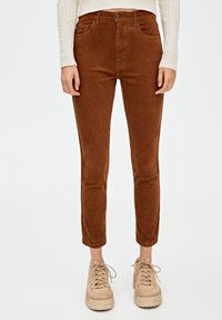 Femme portant un pantalon court en velours côtelé marron, un pull crème à torsades et des baskets à plateforme beige, debout devant un fond blanc.