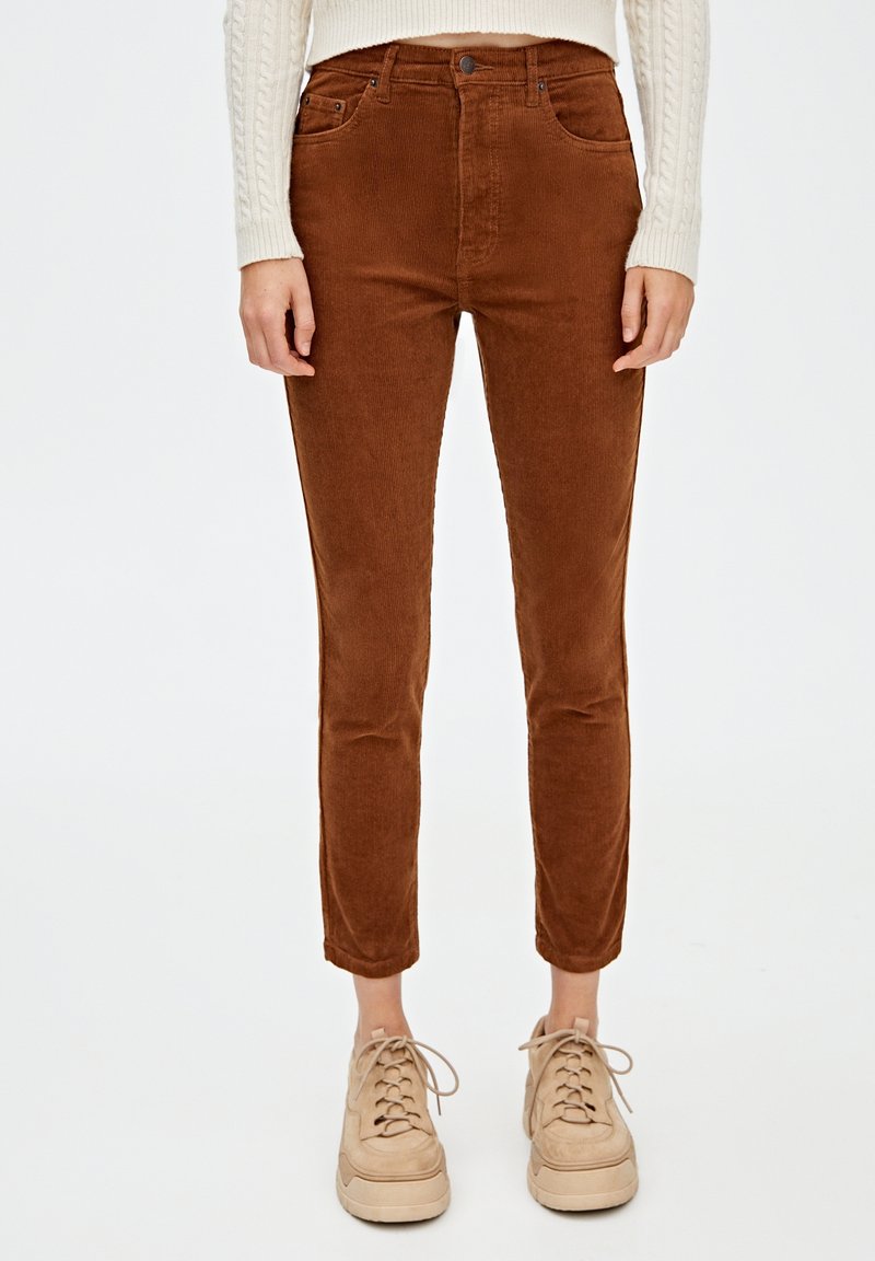 Femme portant un pantalon court en velours côtelé marron, un pull crème à torsades et des baskets à plateforme beige, debout devant un fond blanc.