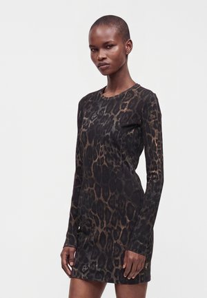 Kvinde med kort hår iført en langærmet, tætsiddende kjole med sort og brun leopardprint, stående mod en ensfarvet lys baggrund.