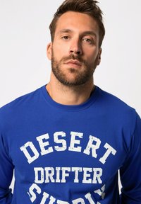 T-shirt a maniche lunghe blu reale realizzato in cotone, con una stampa bianca usurata che recita "DESERT DRIFTER SUPPLY" sul petto.