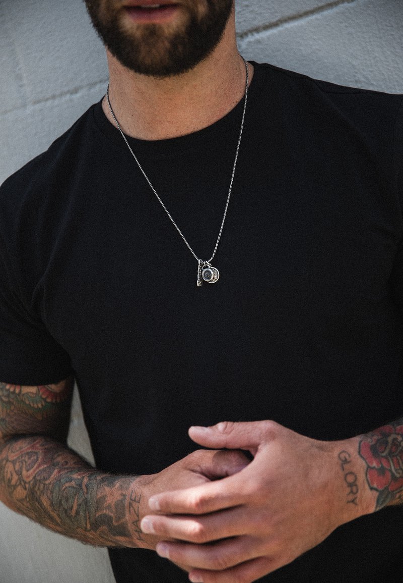 Silberne Halskette mit runden und Anhänger-Charms, über einem schwarzen T-Shirt getragen. Filigrane Tattoo-Details an den Armen sichtbar.