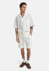 Weißes kurzärmliges Hemd mit strukturiertem Muster, mit Knopfleiste; kombiniert mit heller Shorts und braunen Schnürschuhen.