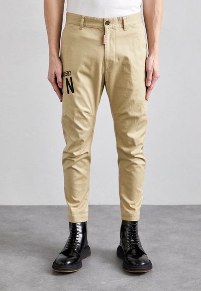 Beige cotton trousers with a slim fit, featuring a black "N" graphic on the left leg, and styled with glossy black lace-up boots.