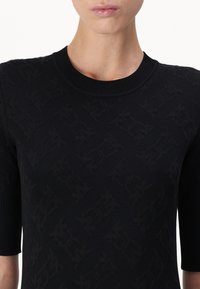 Maglia nera a maglia con scollatura rotonda e maniche corte. Il tessuto presenta un motivo testurizzato di lettere intrecciate, creando un effetto tridimensionale.