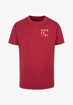 T-shirt en coton rouge avec des manches courtes, un col rond et un graphique en texte blanc affichant "JAMAIS À L'HEURE" à l'avant.