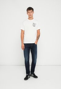 Valge puuvillane T-särk musta trükitud logoga, koos siniste denim-pükstega ja mustade sneakkidega, millel on valged triibud. Mudel seisab valgel taustal.