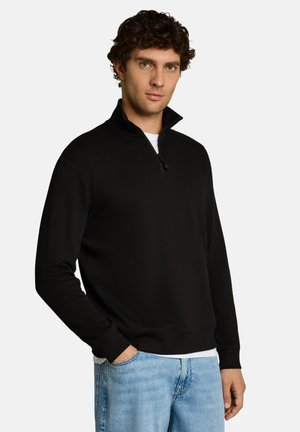 Mann mit lockigem Haar, der einen schwarzen Pullover mit Reißverschluss über einem weißen Hemd und hellblauen Jeans trägt, steht mit einer Hand in der Tasche.