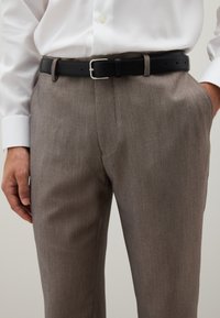 Mann in taupefarbenen Anzughosen mit einem schwarzen Ledergürtel und einem weißen, langärmeligen, in die Hose gesteckten Hemd.