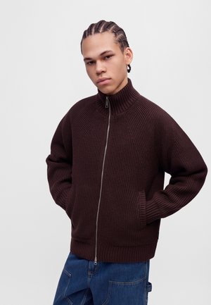 Un modèle aux cheveux tressés pose devant un fond de studio blanc, vêtu d’un pull en maille marron foncé zippé et d’un jean bleu.