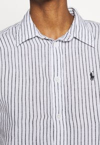 Närbild av en person som bär en vit skjorta med svarta lodräta ränder och en liten svart broderad polo-spelare-logotyp på bröstet.