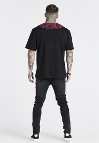 Camiseta negra con estampado de rosas coloridas en la parte superior de la espalda, combinada con jeans oscuros y zapatillas negras. Tejido suave y corte relajado.