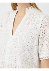 Vit bomulls klänning med intrikata broderie anglaise-mönster. Har en grund V-hals och vågig krage. Korta puffärmar.