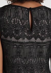 Haut en dentelle noire avec un motif texturé, présentant une fermeture au dos en forme de goutte et un col lisse. Détails complexes sur l'ensemble.