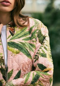 Gestepptes Jacket mit einem rosa Stoffgrund und grünen Blattmustern sowie Schmetterlingsdetails; enthält einen silbernen Reißverschluss und einen gerippten Kragen.