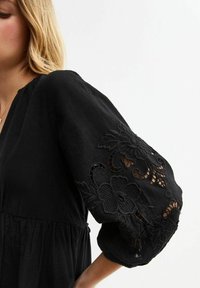 Robe noire à coupe ample avec un motif floral brodé de manière complexe sur les manches. Fabriquée dans un tissu texturé avec des manches à volants généreuses.