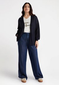 Marinefarbener Cardigan mit strukturierten Ärmeln über einem weißen T-Shirt mit glitzerndem Text, kombiniert mit weit geschnittenen Jeans und beigen Sneakers.