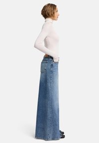 Gonna maxi in denim con una leggera svasatura, caratterizzata da un lavaggio medio, tasche posteriori e un design tradizionale a cinque tasche. Indossata con un dolcevita aderente.