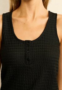 Black tank top with a textured fabric, featuring a round neck and three black button accents at the front.