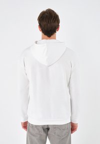 White cotton hoodie with a relaxed fit, featuring a drawstring hood, long sleeves, and a smooth texture. Gray pants visible below.