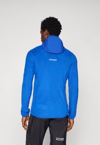 Μπλε φούτερ με μακριά μανίκια, με υφασμάτινο σχέδιο, κουκούλα και λογότυπο στο πίσω μέρος. Συνδυασμένο με μαύρο παντελόνι που διαθέτει λογότυπο της μάρκας.