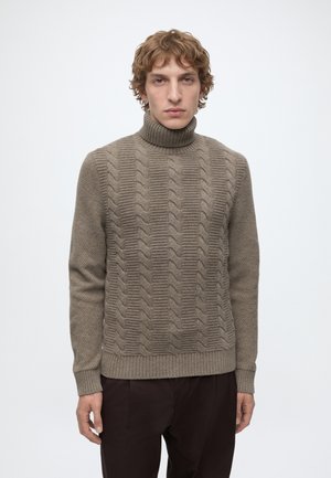 Man met kort krullend haar die een bruine kabelgebreide coltrui en donkere broek draagt, tegen een effen lichte achtergrond.