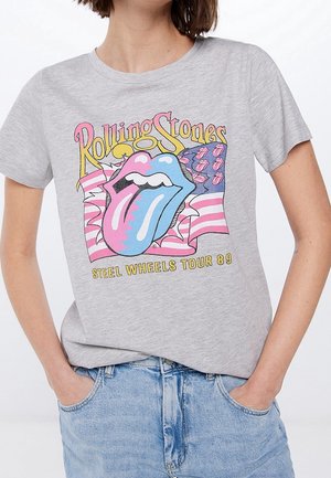 Persona che indossa una maglietta grigia del Rolling Stones Steel Wheels Tour 89 con una grafica colorata della lingua e della bandiera, abbinata a jeans azzurri chiari.