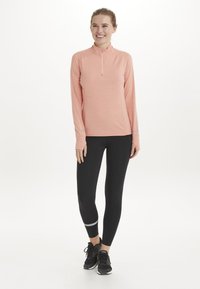 Langærmet zip-neck top i blød koralfarvet stof, parret med sorte leggings med reflekterende detaljer og sorte træningssko.