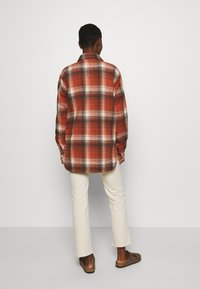 Rood en zwart geruite flanellen blouse met kraag, lange mouwen en een rechte snit, gedragen over lichtgekleurde broek en bruine sandalen.