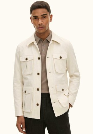 Mann trägt eine cremefarbene, geknöpfte Jacke mit vier vorderen Taschen über einem taupefarbenen Poloshirt und schwarzen Hosen, steht mit einer Hand in der Tasche.
