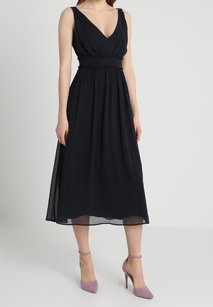Donna che indossa un abito midi nero senza maniche con scollo a V e vita arricciata, abbinato a scarpe con tacchi alti color lilla chiaro.