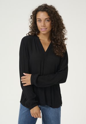 Blouse noire à manches longues avec un col en V et un tissu texturé, dotée de poignets froncés ; assortie à un jean en denim bleu clair.