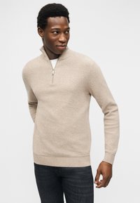 Άνδρας που φοράει μπεζ πουλόβερ με φερμουάρ μέχρι το στήθος και σκούρο τζιν, στέκεται με απαλά χαλαρωμένα τα χέρια, κοιτάζοντας ελαφρώς προς τα αριστερά του, μπροστά σε απλό λευκό φόντο.