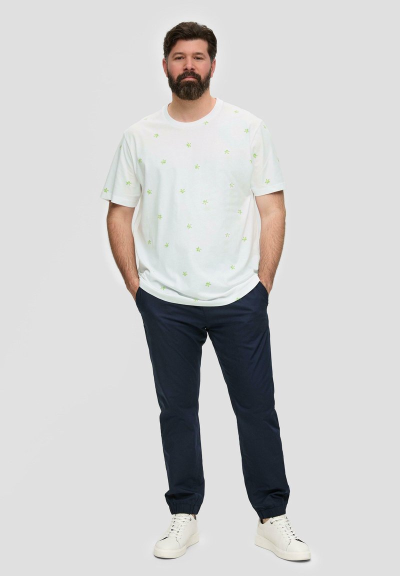 Witte t-shirt met korte mouwen en groene bloemenpatronen, gecombineerd met donkerblauwe broek en witte sneakers. Eenvoudig ontwerp en een ontspannen pasvorm.