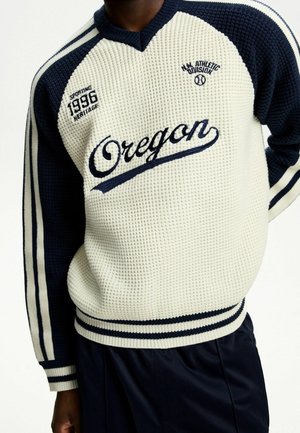 Personne portant un pull en maille gaufrée crème et bleu marine avec le texte "Oregon", des manches rayées bleu marine, et un lettrage de style athlétique sur la poitrine.