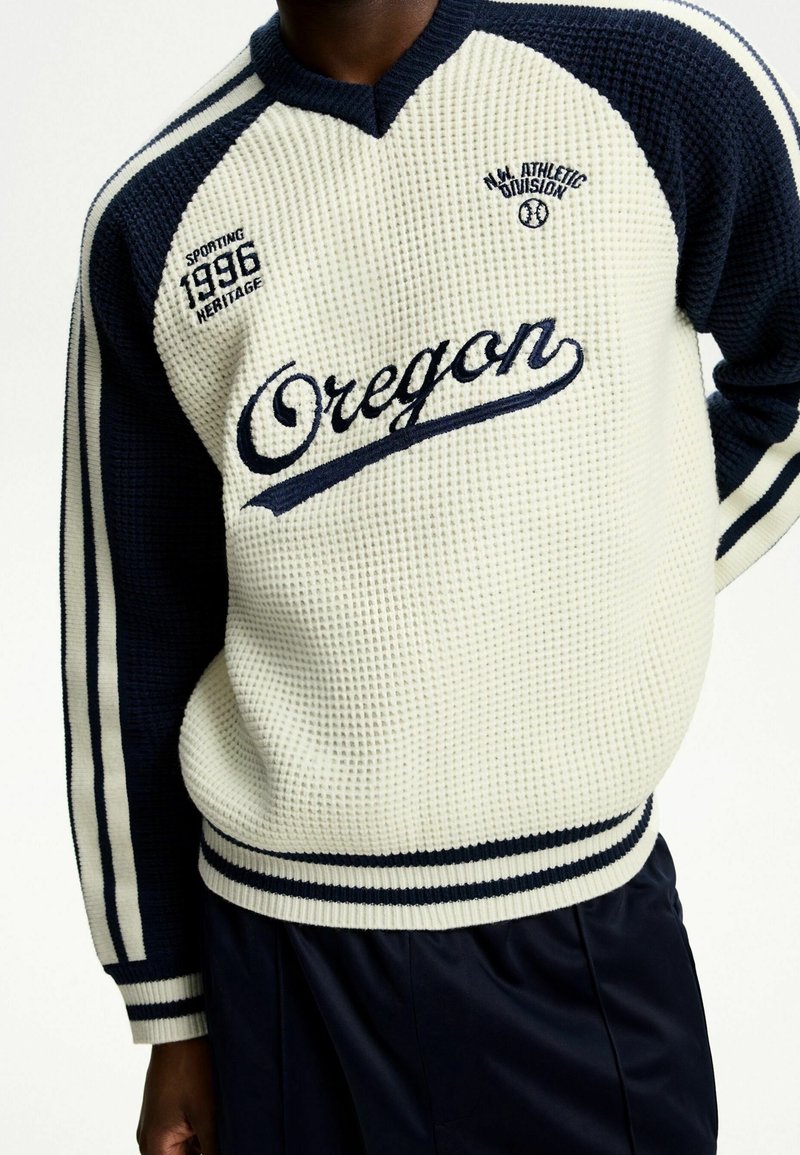 Personne portant un pull en maille gaufrée crème et bleu marine avec le texte "Oregon", des manches rayées bleu marine, et un lettrage de style athlétique sur la poitrine.